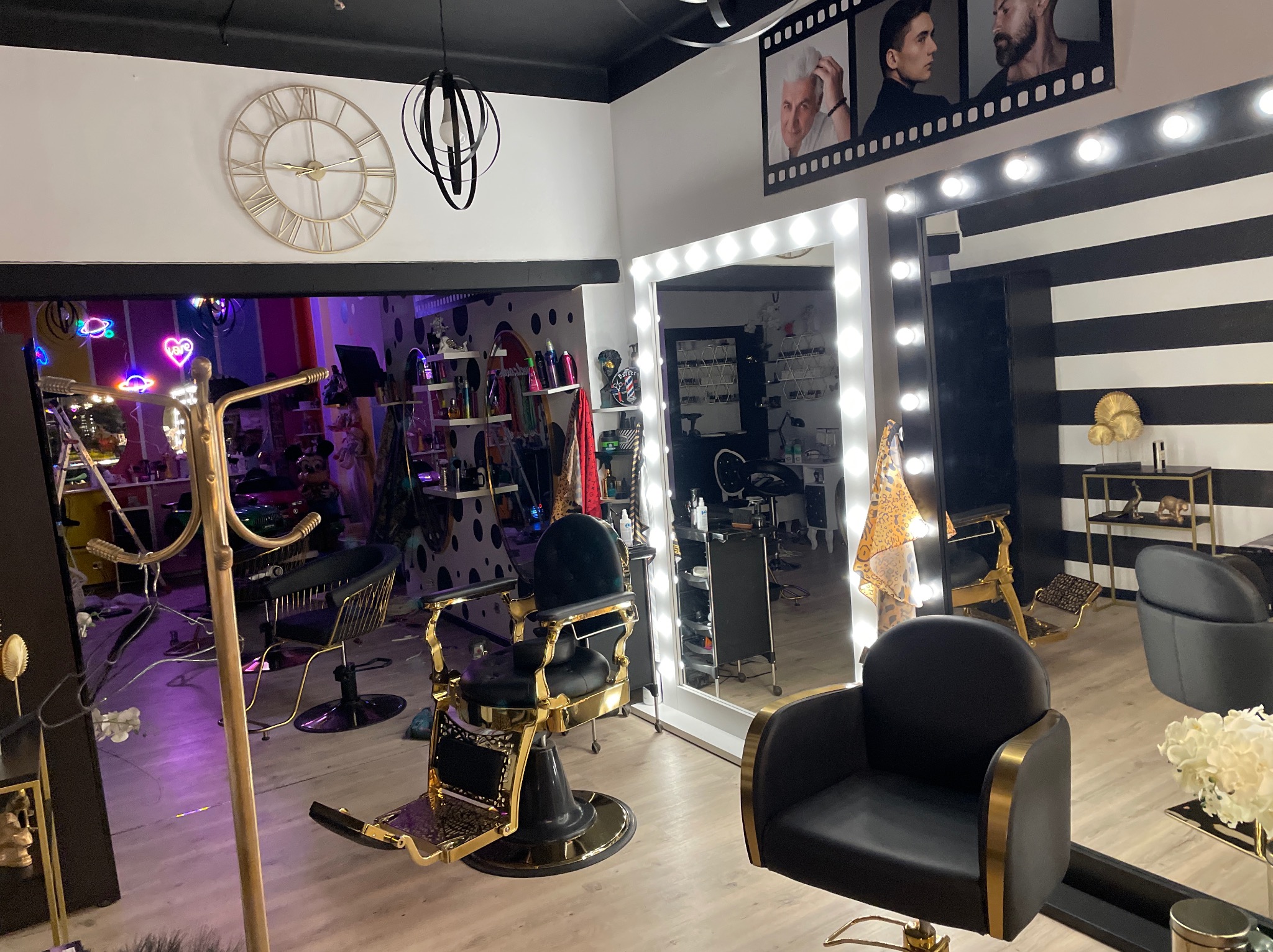 Wnętrze salonu fryzjerskiego z dwoma fotelami, lustrem z oświetleniem i dekoracjami ściennymi. Widoczny zegar i neonowe ozdoby.