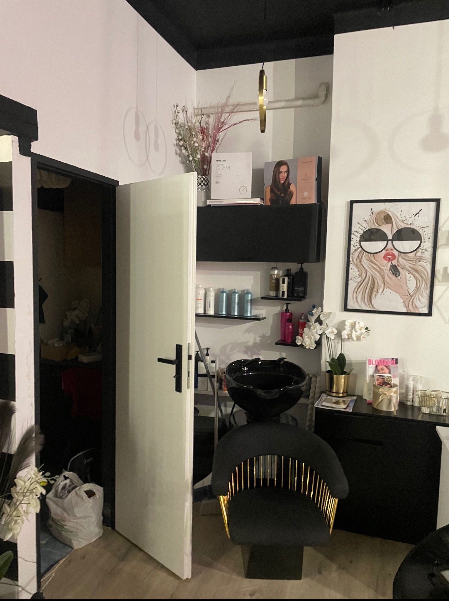 Nowoczesne wnętrze salonu fryzjerskiego z czarnym fotelem, umywalką, półkami z kosmetykami i plakatem z wizerunkiem kobiety w okularach. Widoczne otwarte drzwi.