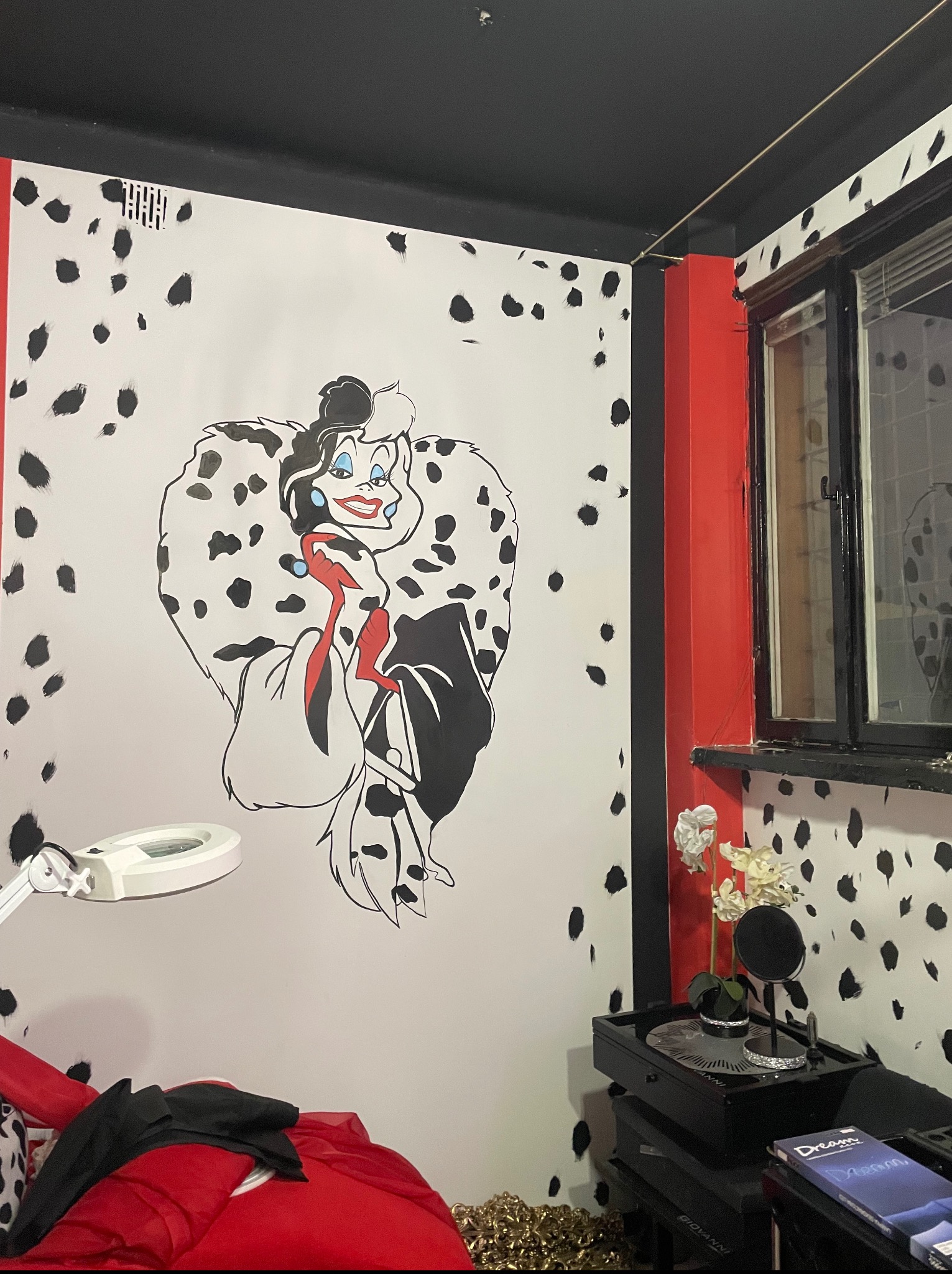 Pokój z motywem Cruelli De Mon: białe ściany w czarne kropki, czerwony akcent przy oknie i duży rysunek postaci. Widoczna lampa powiększająca i dekoracje.