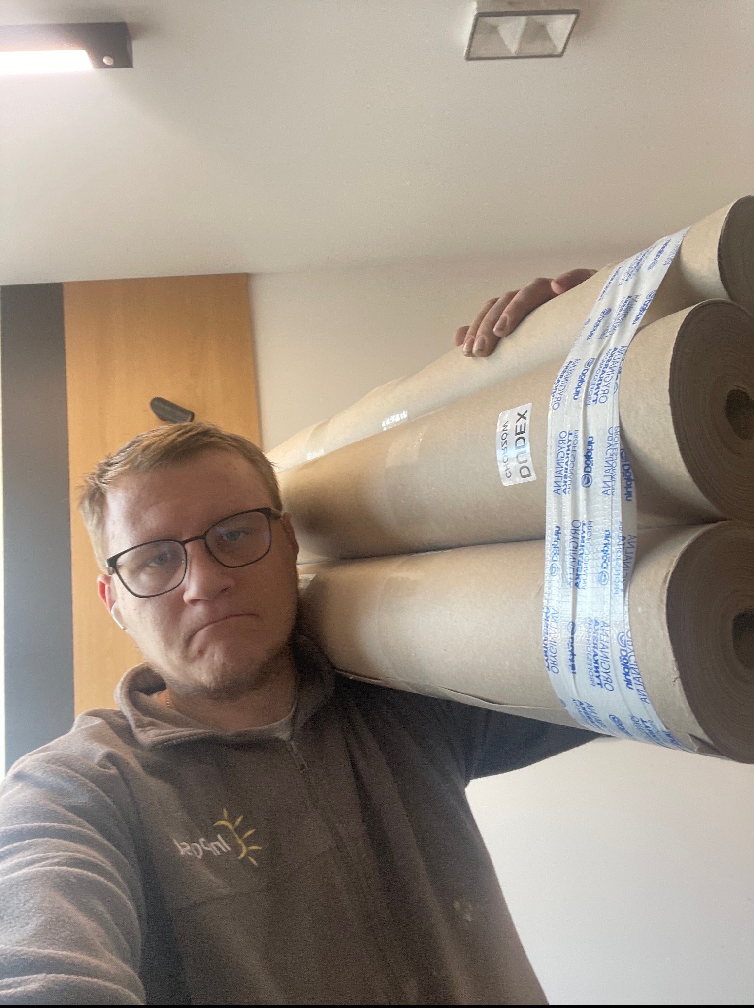 Mężczyzna w okularach niesie na ramieniu kilka rolek materiału budowlanego, prawdopodobnie do wykończenia wnętrz. Widoczna etykieta 'DODEX'.
