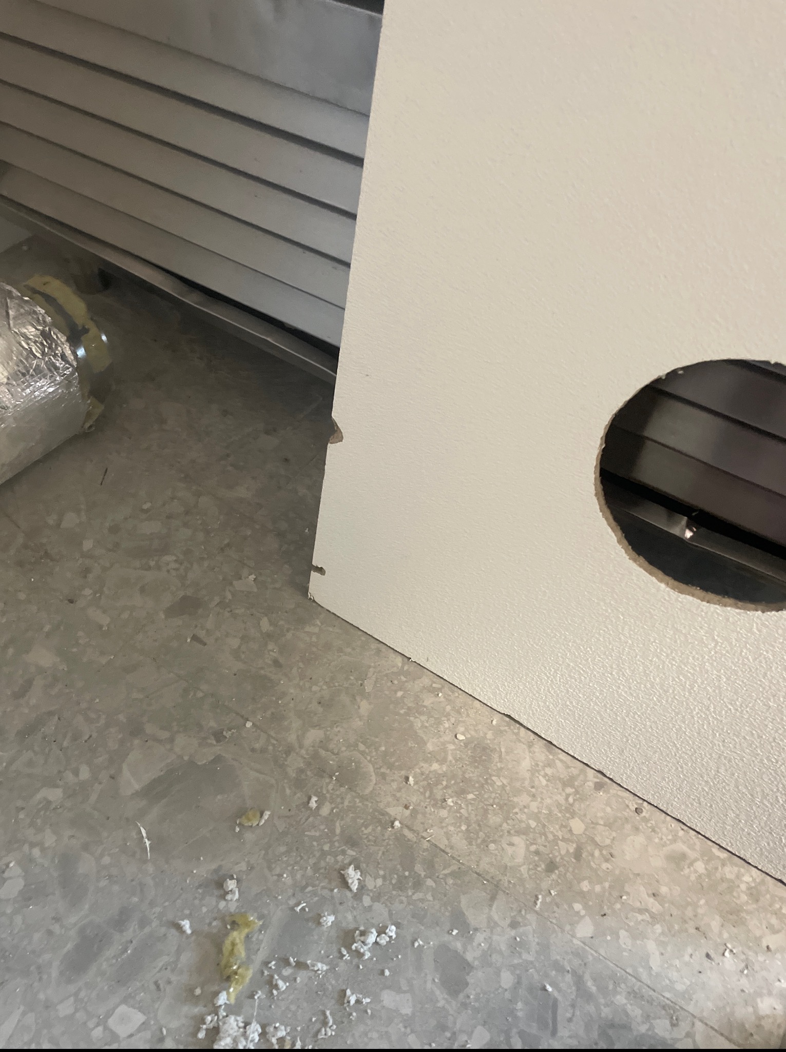 Białe drzwi z wyciętym okrągłym otworem, widoczna kratka wentylacyjna. Na podłodze resztki materiałów budowlanych. W tle kratka wentylacyjna.