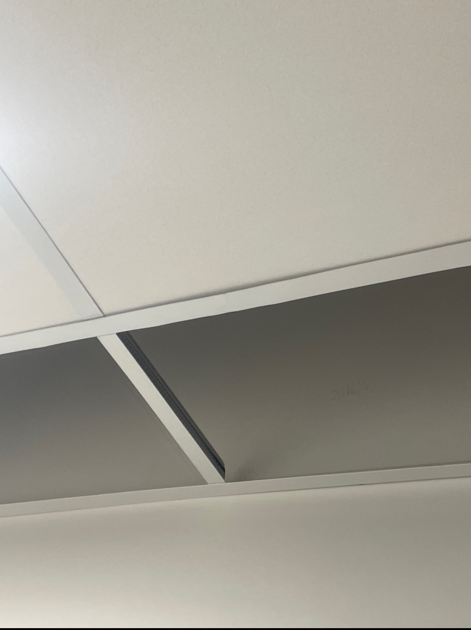 Zamontowany sufit podwieszany z kasetonami, widok z dołu. Białe profile i szare panele tworzą geometryczny wzór. Widoczne niedoskonałości na panelu.