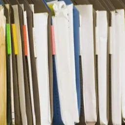 Rząd segregatorów z dokumentami, z kolorowymi etykietami indeksującymi, ustawionych pionowo w szafie biurowej. Widok z boku, lekko z góry.