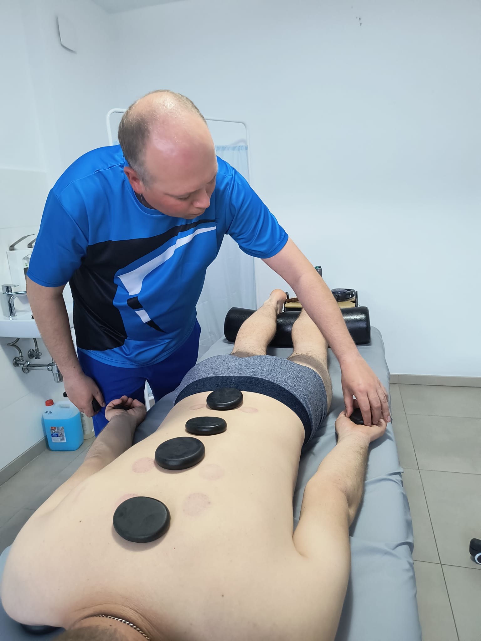 Terapeuta wykonuje zabieg gorącymi kamieniami na plecach klienta leżącego na stole do masażu. Na skórze widoczne ślady po poprzednich zabiegach.
