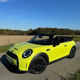 Wiktoria Konica - Żółty Mini Cooper kabriolet z czarnymi felgami stoi na asfalcie, w tle pole i las pod błękitnym niebem. Auto sfotografowane z boku, w słoneczny dzień.