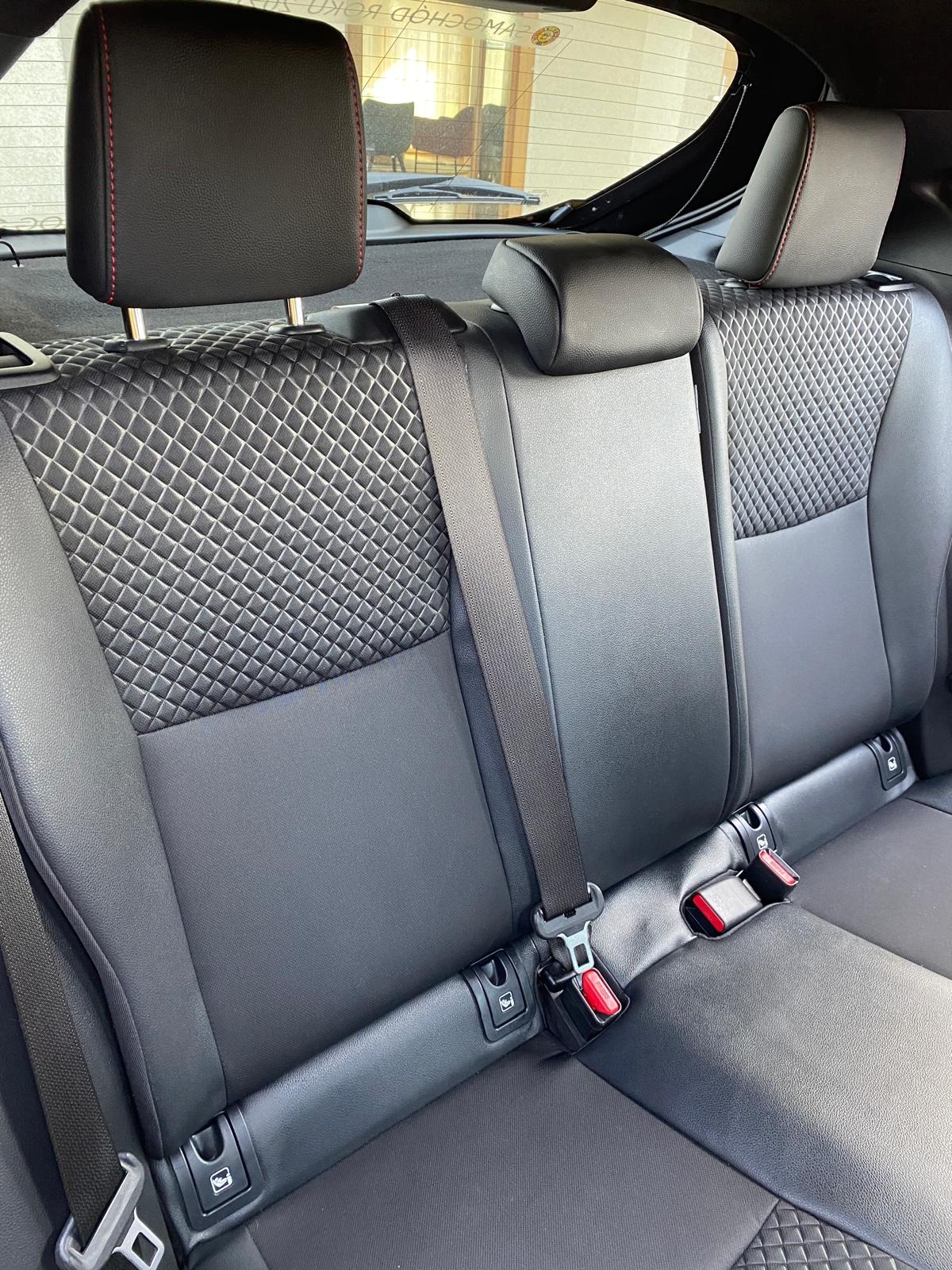 Czarne, pikowane tylne siedzenia samochodu z pasami bezpieczeństwa. Widoczny zagłówek z czerwoną nicią. W tle szyba i odbicie wnętrza budynku.