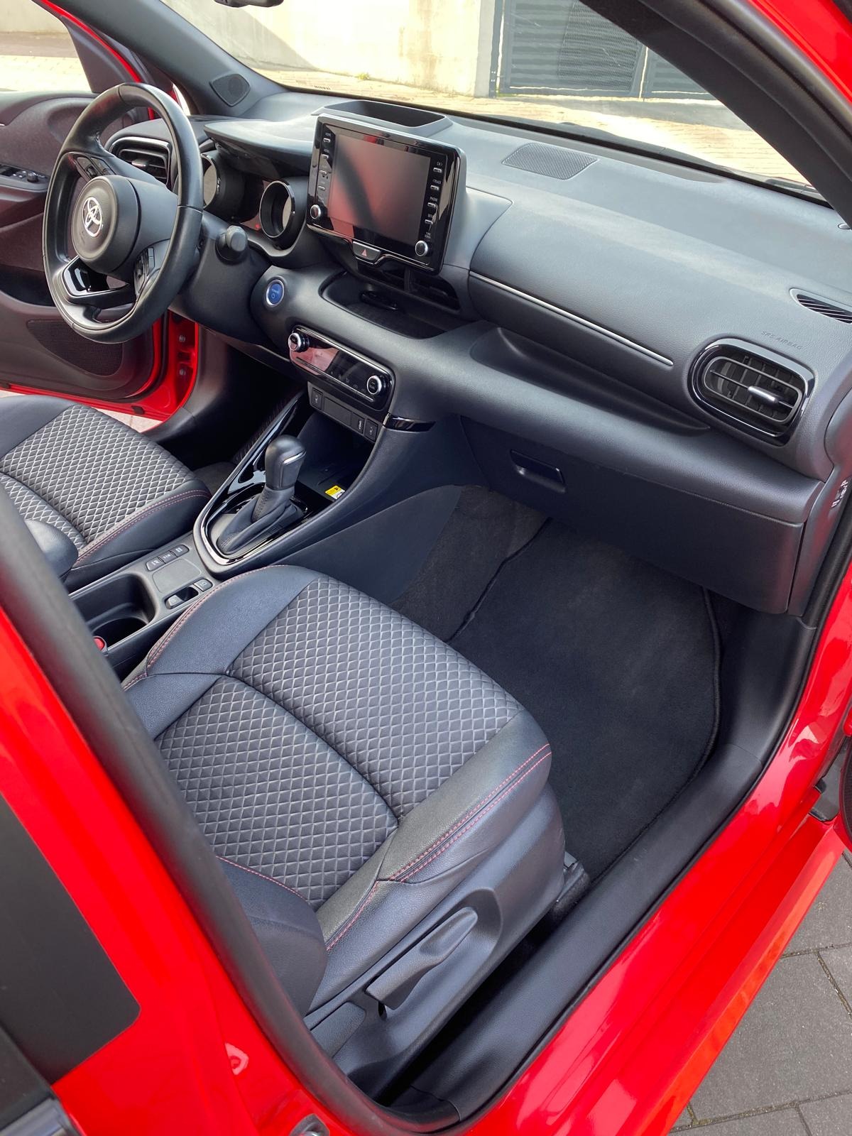 Wnętrze czerwonego samochodu, widok na deskę rozdzielczą, fotele z geometrycznym wzorem i czarne dywaniki. Detale wykończenia, ergonomiczny design.