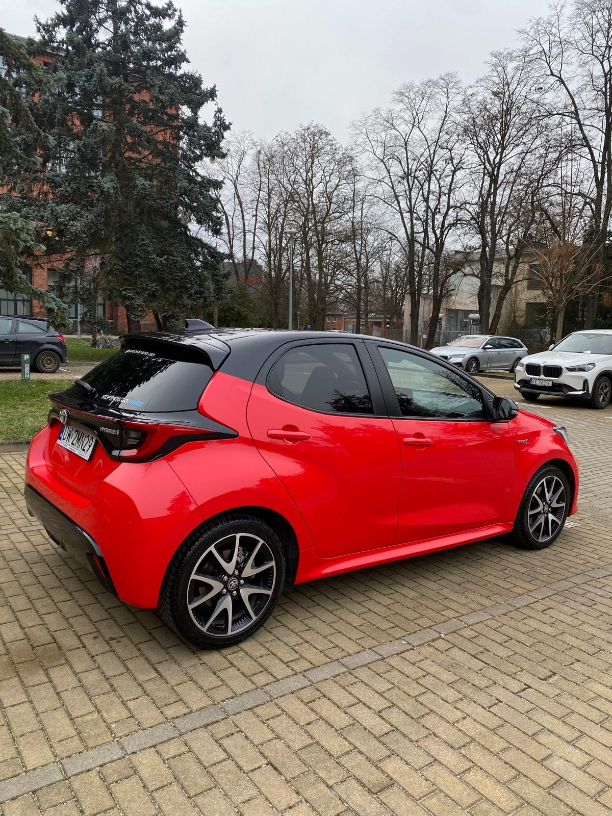 Czerwony, miejski hatchback Toyota Yaris Hybrid z czarnym dachem, zaparkowany na brukowanym placu, z widocznymi innymi autami i drzewami w tle.