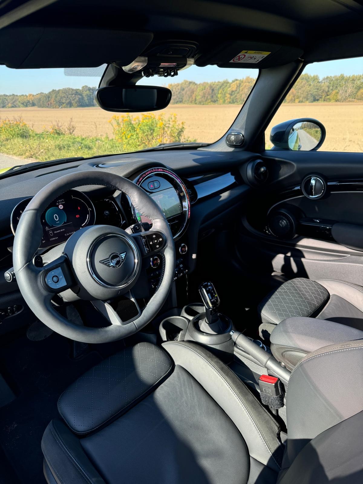 Wnętrze nowoczesnego samochodu Mini Cooper z widokiem na kierownicę, deskę rozdzielczą i skórzane fotele. Za szybą krajobraz polny w słoneczny dzień.