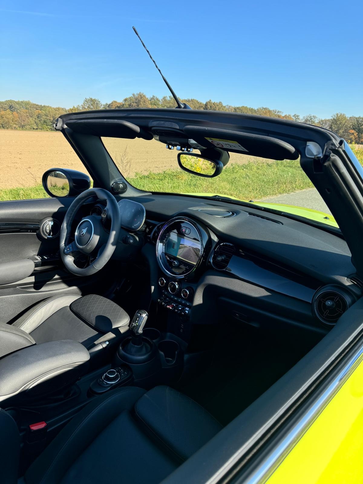 Wnętrze kabrioletu Mini Cooper z czarną tapicerką skórzaną i widokiem na krajobraz za oknem. Jasnozielony lakier karoserii i nowoczesny design deski rozdzielczej.