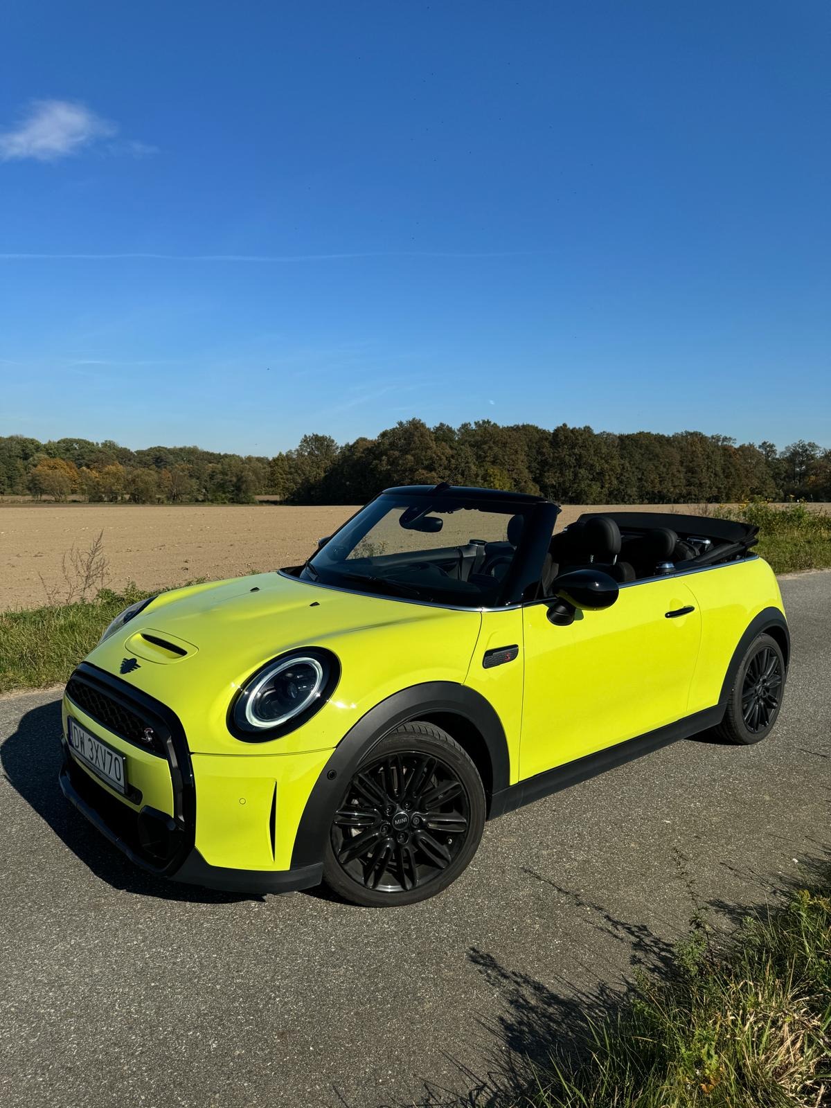 Żółty Mini Cooper kabriolet z czarnymi felgami stoi na asfalcie, w tle pole i las pod błękitnym niebem. Auto sfotografowane z boku, w słoneczny dzień.
