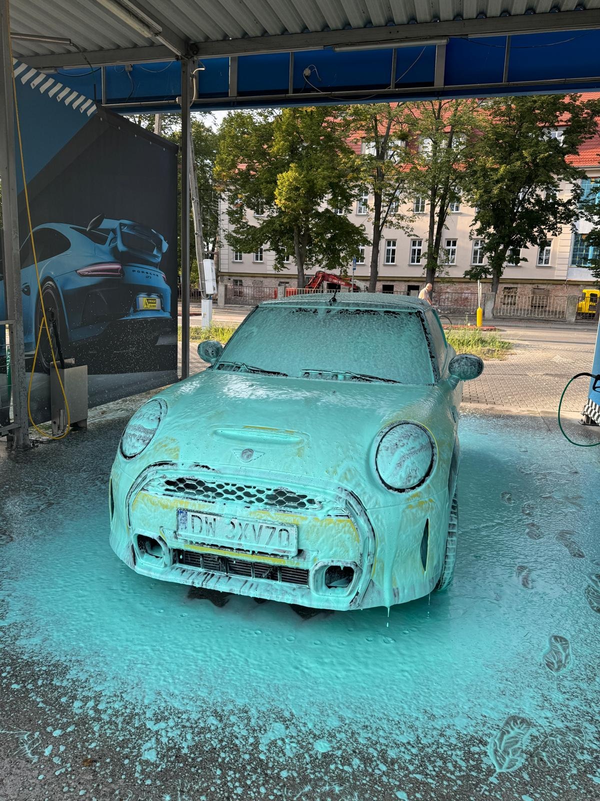 Mini Cooper pokryty pianą podczas mycia w myjni samochodowej z plakatem Porsche w tle, widziany z przodu na zewnątrz.