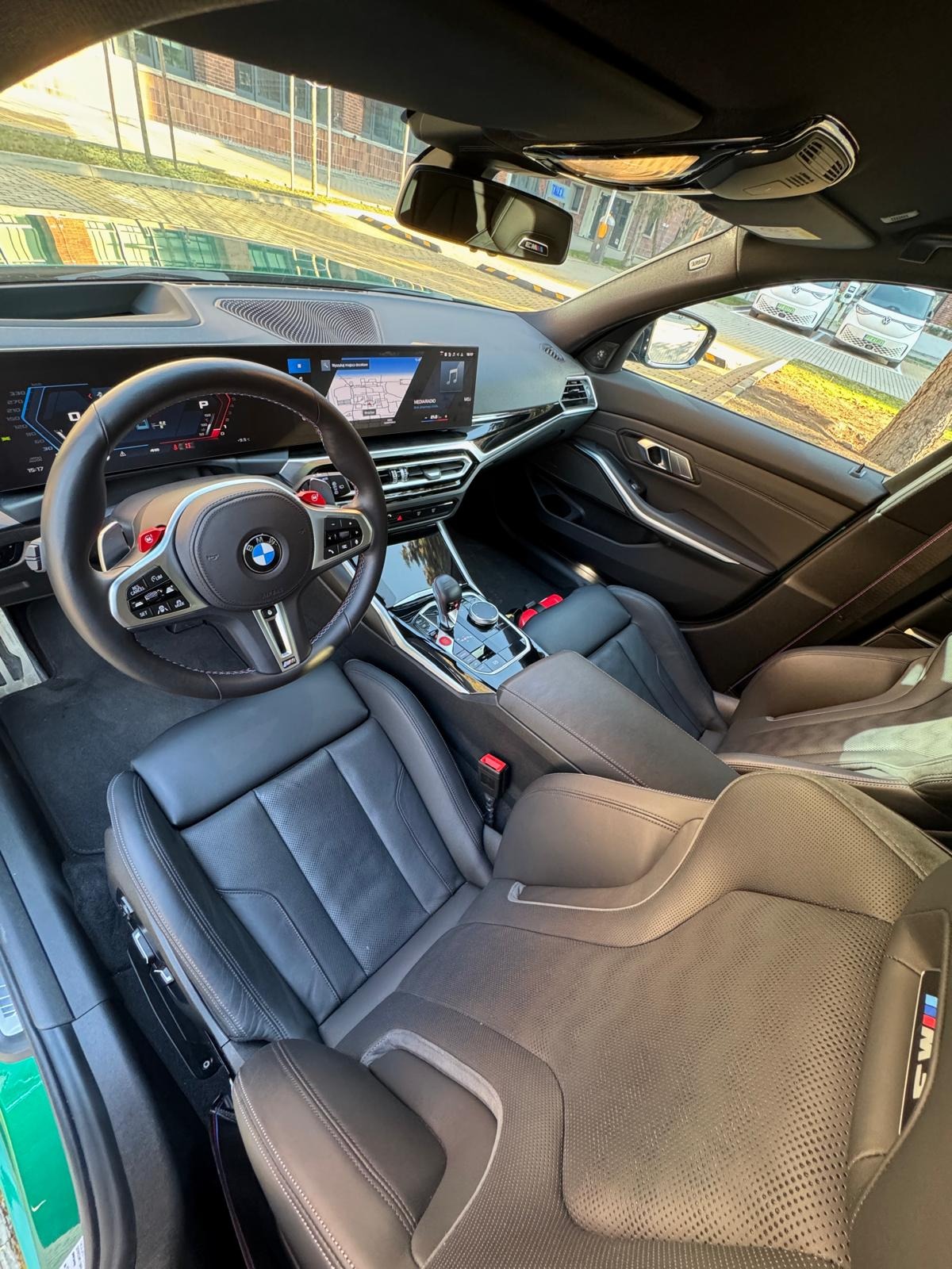 Wnętrze luksusowego BMW M3 w zielonym kolorze z czarną skórzaną tapicerką. Widoczne fotele kubełkowe, deska rozdzielcza z dużym ekranem i sportowa kierownica.