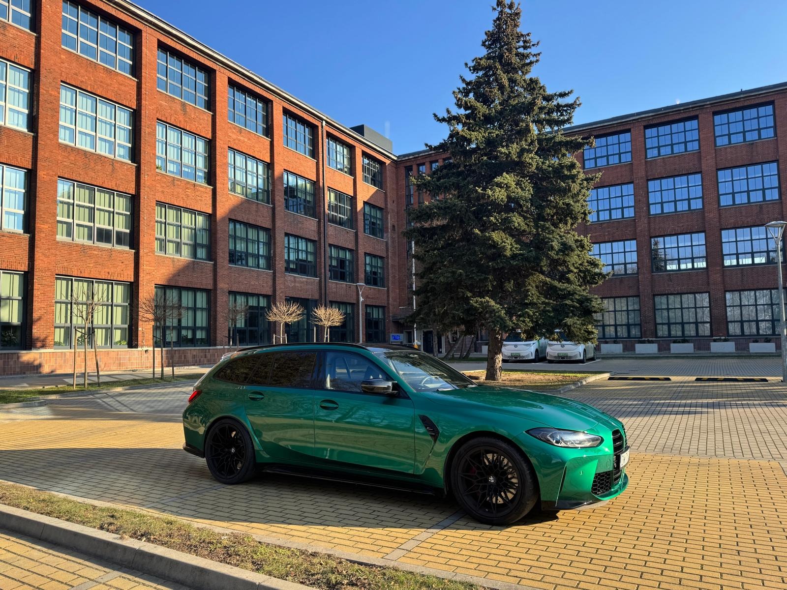 Zielone BMW M3 Touring na tle ceglanego budynku z dużymi oknami i zielonego drzewa. Samochód stoi na żółtej kostce brukowej w słoneczny dzień.
