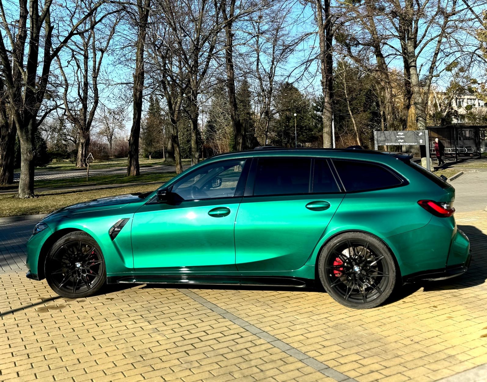 Szmaragdowy, metaliczny samochód kombi z czarnymi felgami i czerwonymi zaciskami hamulcowymi, zaparkowany na brukowanym placu w słoneczny dzień.