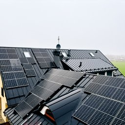 Innowacyjny Dom - Instalacja fotowoltaiczna na dachu domu jednorodzinnego z czarną blachodachówką, widok z góry w pochmurny dzień. W tle zielone pole.