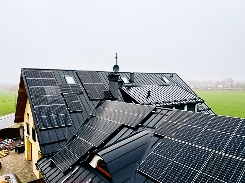 Instalacja fotowoltaiczna na dachu domu jednorodzinnego z czarną blachodachówką, widok z góry w pochmurny dzień. W tle zielone pole.