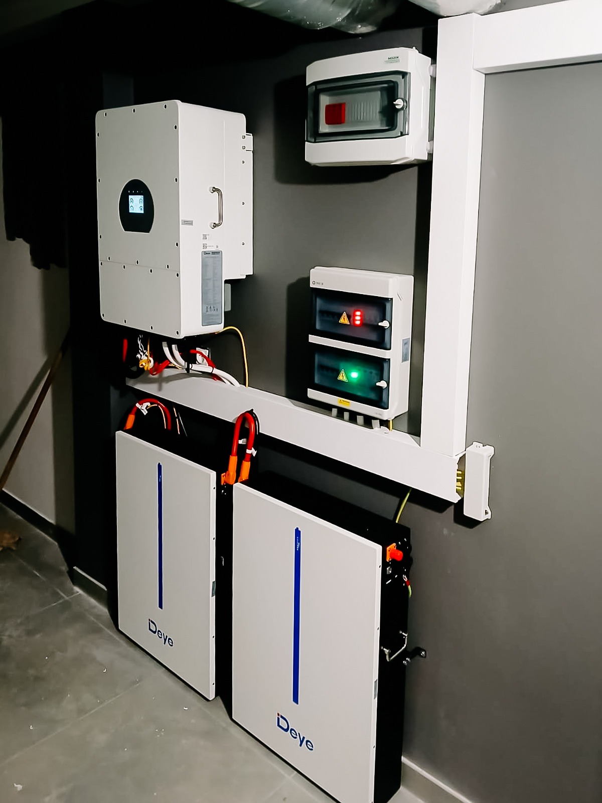 Kompletna instalacja magazynu energii Deye z inwerterem i zabezpieczeniami. Widoczne okablowanie i podłączenia. Szara ściana, betonowa podłoga.