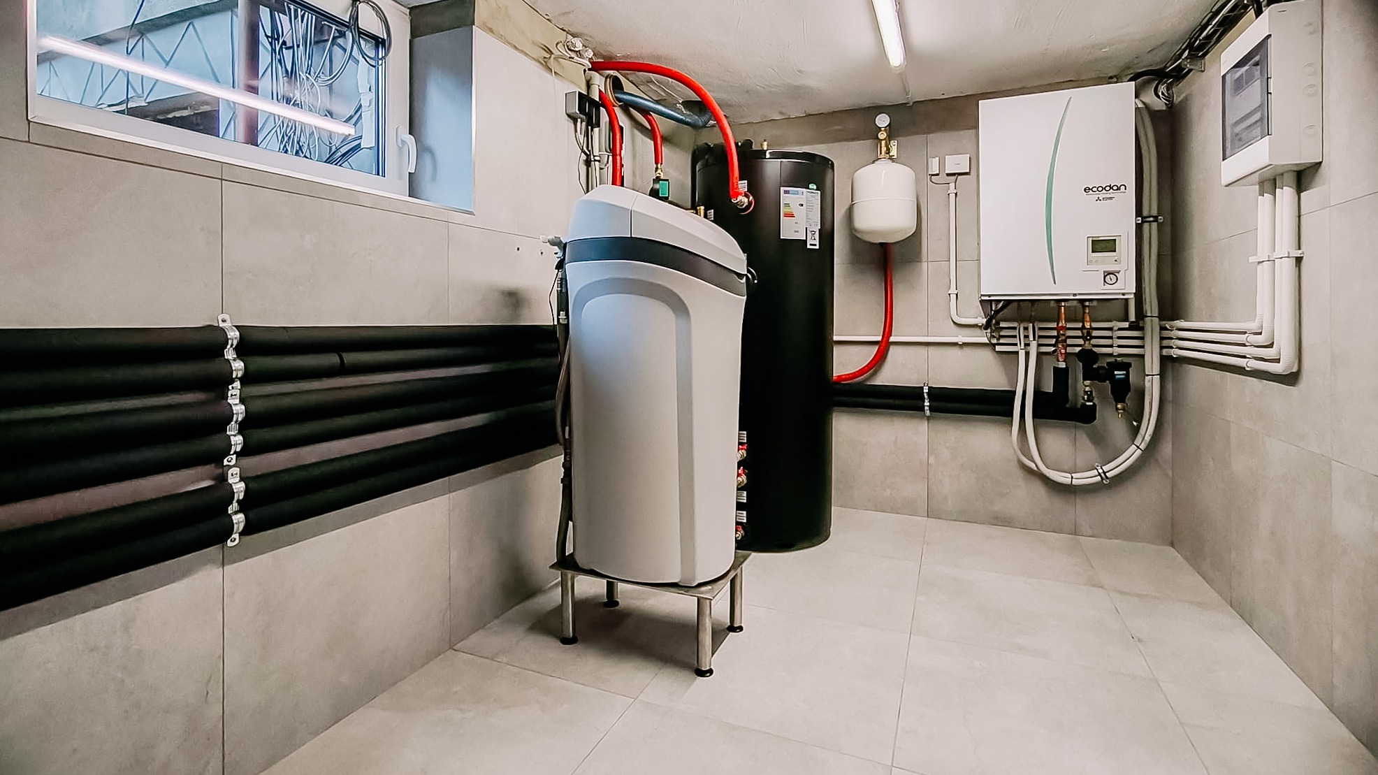 Zestaw instalacji grzewczej w kotłowni: zmiękczacz wody na podeście, zasobnik CWU, pompa ciepła Ecodan. Rury w izolacji, szare płytki ścienne i podłogowe.