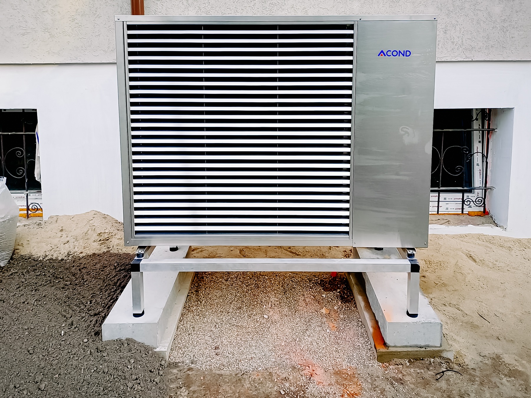 Zewnętrzna jednostka pompy ciepła Acond, zamontowana na betonowej podstawie, z widocznymi oknami piwnicznymi w tle. Detal instalacji grzewczej.