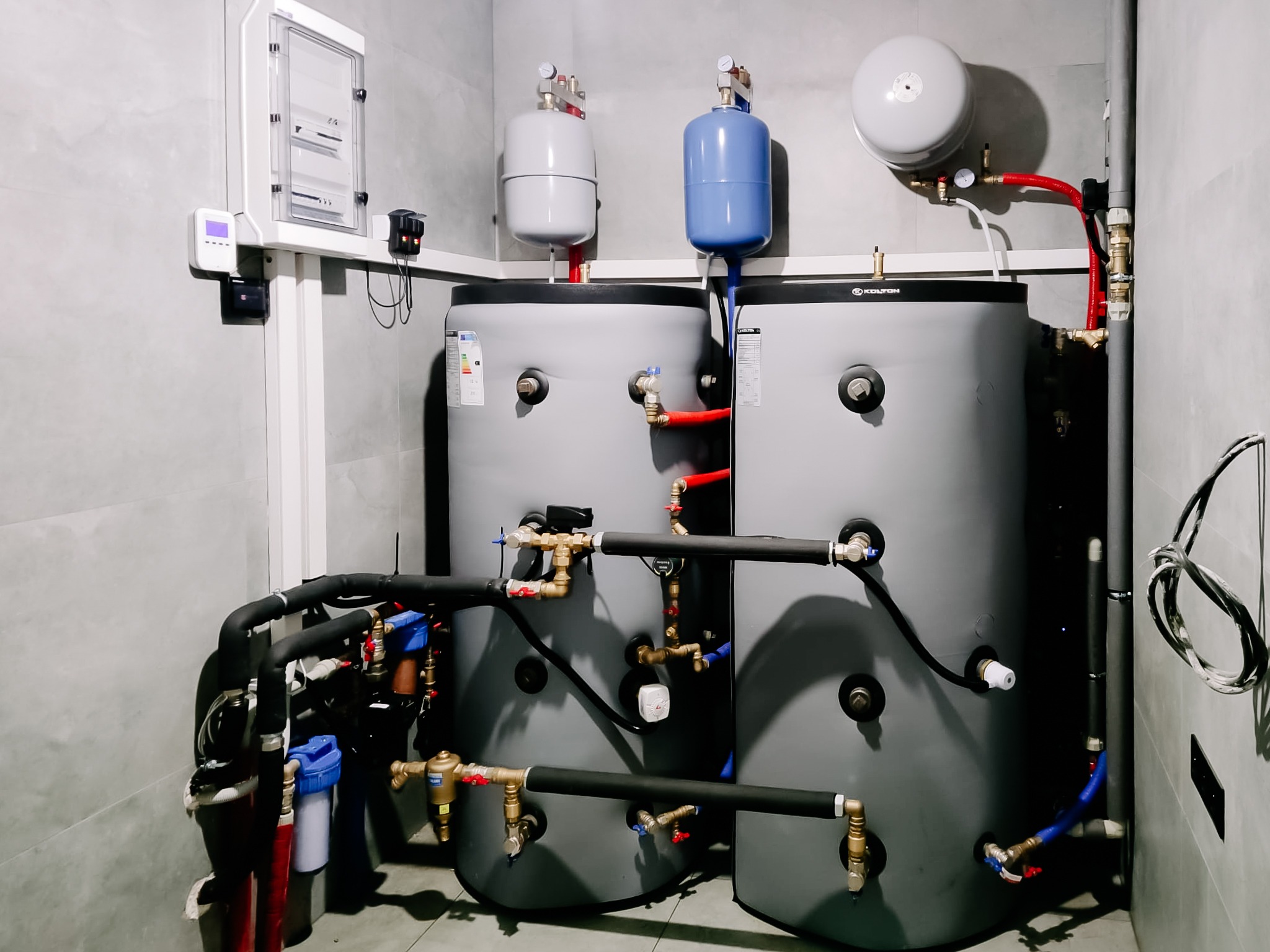 Kotłownia z dwoma szarymi zbiornikami, rurami i osprzętem. Widoczna instalacja wodna i elektryczna. Szare płytki na ścianach.