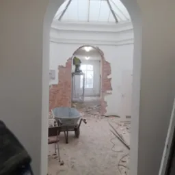 Pracownik na rusztowaniu w trakcie usuwania fragmentu ceglanej ściany w pomieszczeniu z przeszklonym dachem, widok przez łukowate przejście, gruz na podłodze, taczka obok krzesła.