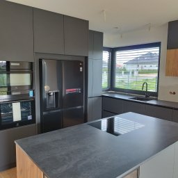 Zakład Stolarski ADB Jarosław Mazur - Nowoczesna, grafitowa kuchnia z wyspą, sprzętem AGD marki Electrolux i LG, oraz drewnianymi akcentami w zabudowie. Widok na okno z żaluzjami i dom za nim.