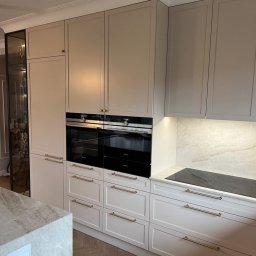 Zakład Stolarski ADB Jarosław Mazur - Jasna, elegancka kuchnia na wymiar z frezowanymi frontami, złotymi uchwytami i zabudowanym sprzętem AGD. Widoczna przeszklona witryna.