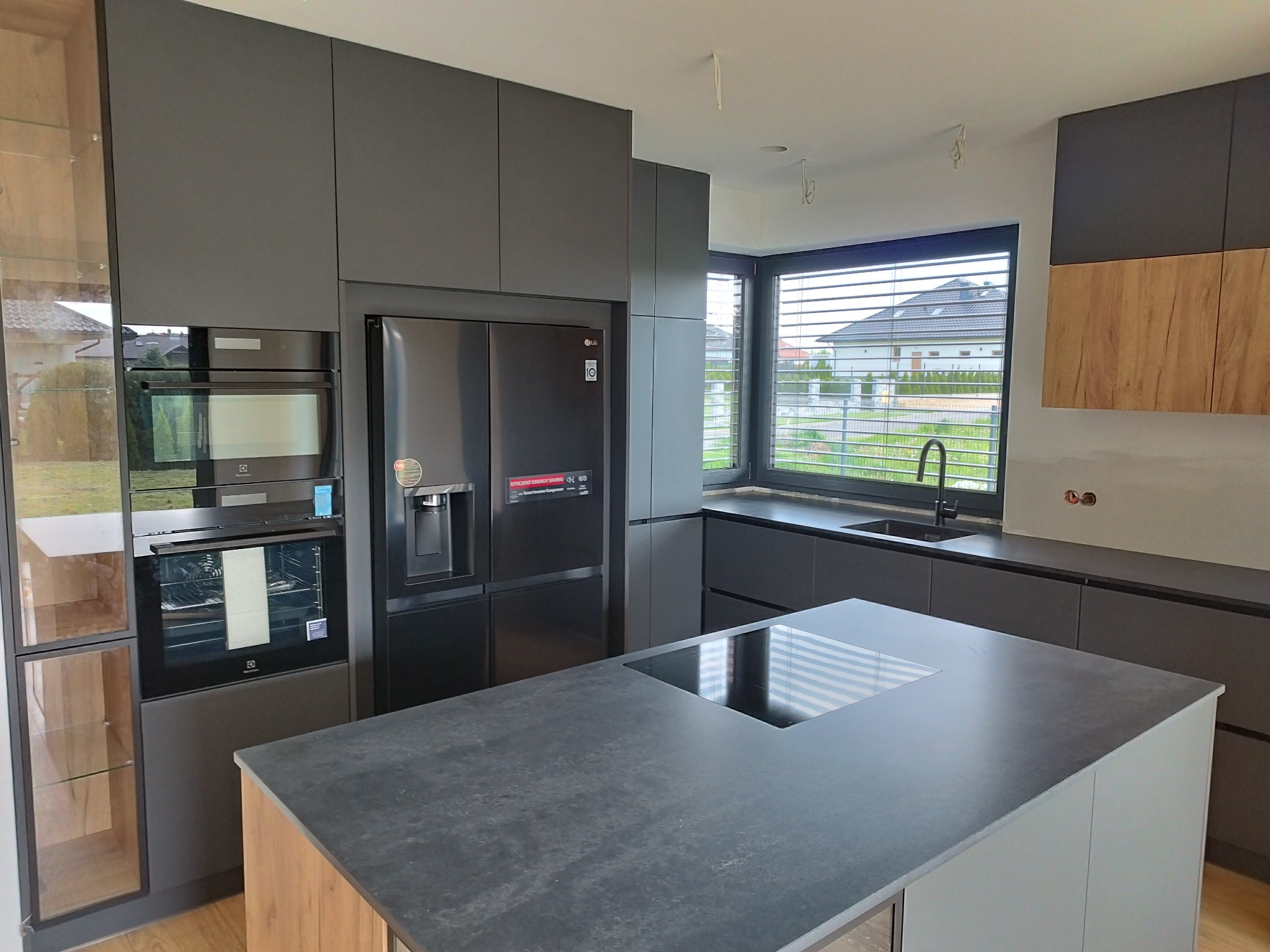 Nowoczesna, grafitowa kuchnia z wyspą, sprzętem AGD marki Electrolux i LG, oraz drewnianymi akcentami w zabudowie. Widok na okno z żaluzjami i dom za nim.