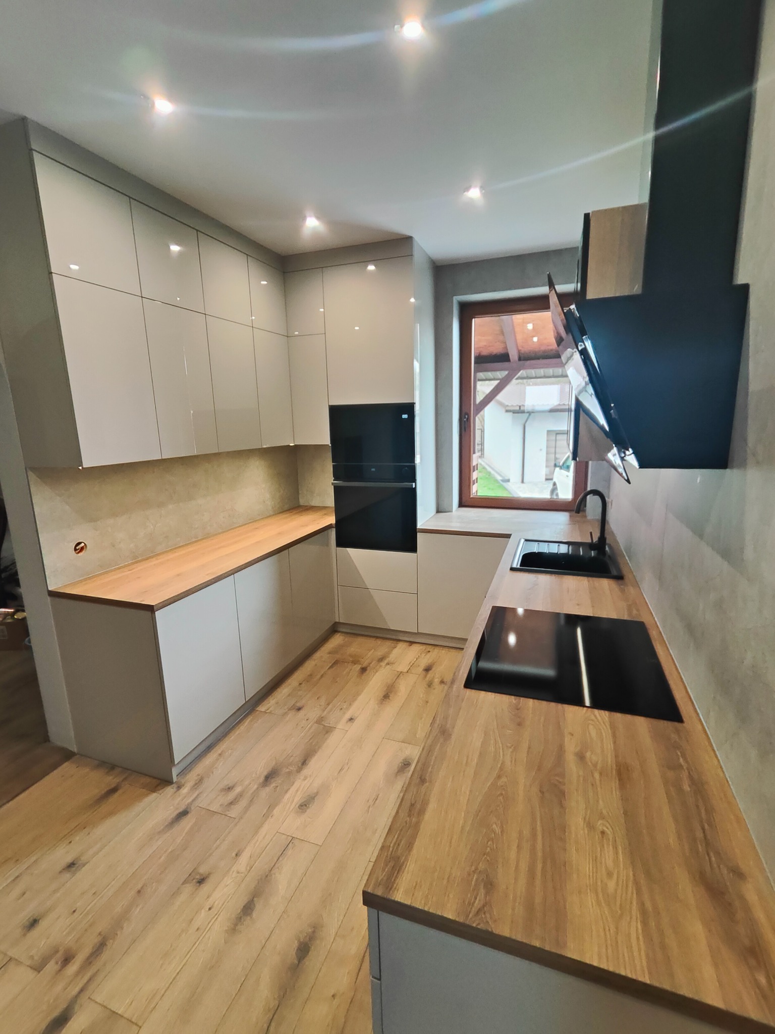 Nowoczesna kuchnia z białymi szafkami i drewnianym blatem, czarnym zlewem i płytą indukcyjną. Minimalistyczny design, jasne wnętrze z oknem i drewnianą podłogą.