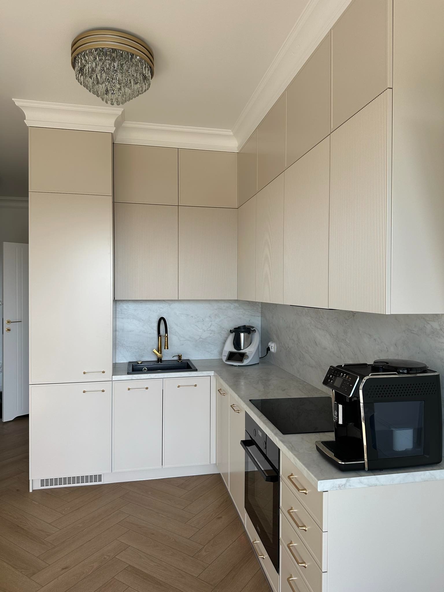 Jasna kuchnia na wymiar z frezowanymi frontami szafek górnych, marmurowym blatem i parkietem w jodełkę. Złota bateria i kryształowy żyrandol dodają elegancji.