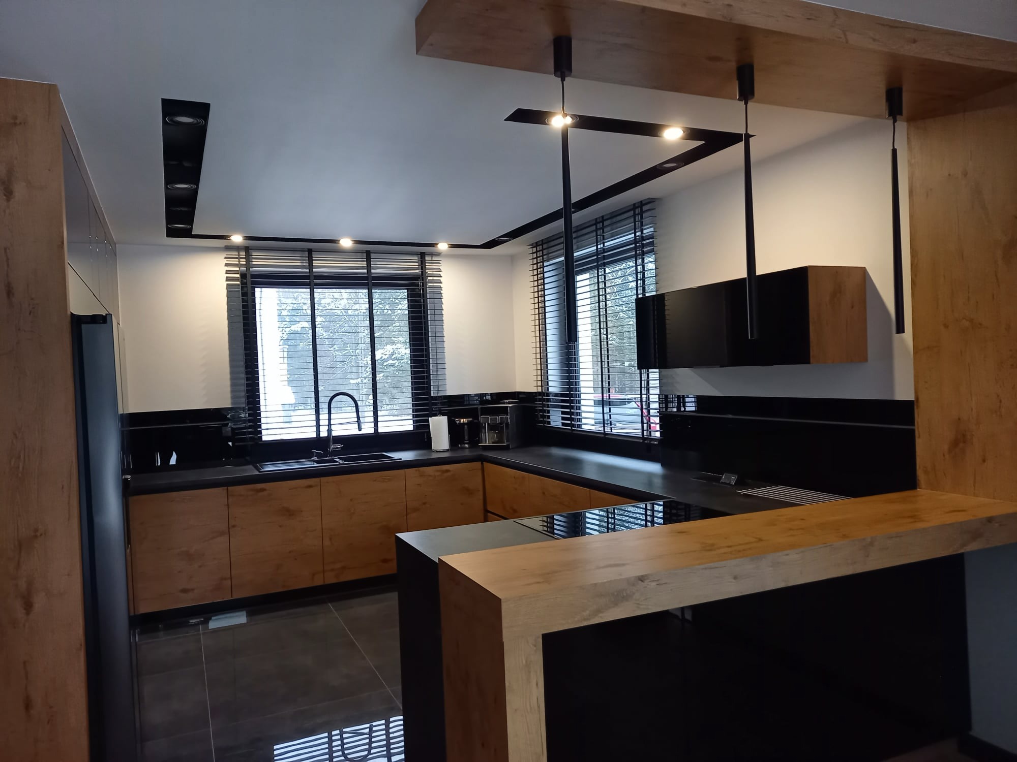 Nowoczesna kuchnia z drewnianymi frontami, czarnym blatem i oświetleniem punktowym. Widoczne okno z żaluzjami i elementy dekoracyjne. Ujęcie z perspektywy użytkownika.