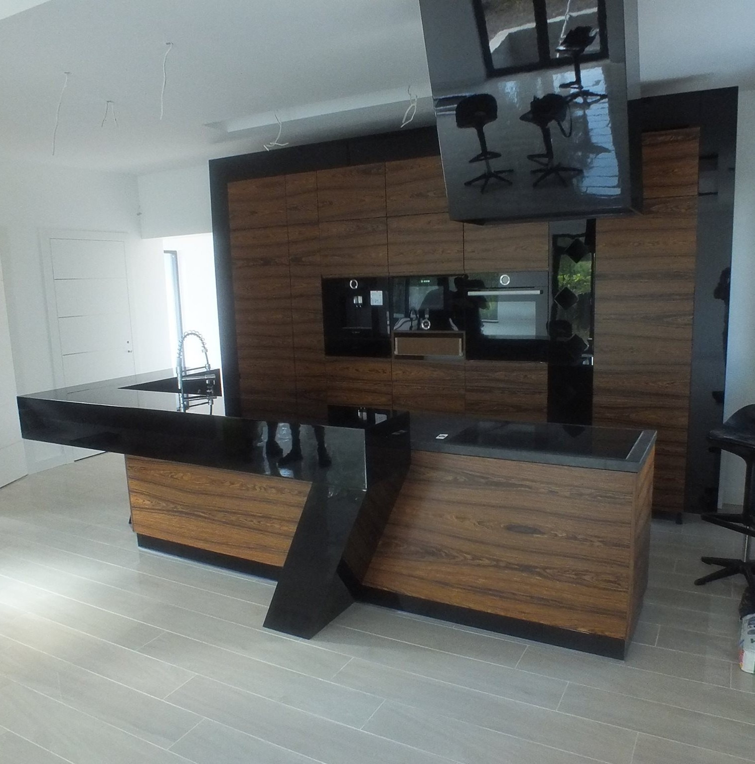 Nowoczesna kuchnia z wyspą: drewniane fronty szafek z czarnymi, błyszczącymi blatami. Minimalistyczny design, oświetlenie punktowe, płytki podłogowe imitujące drewno.