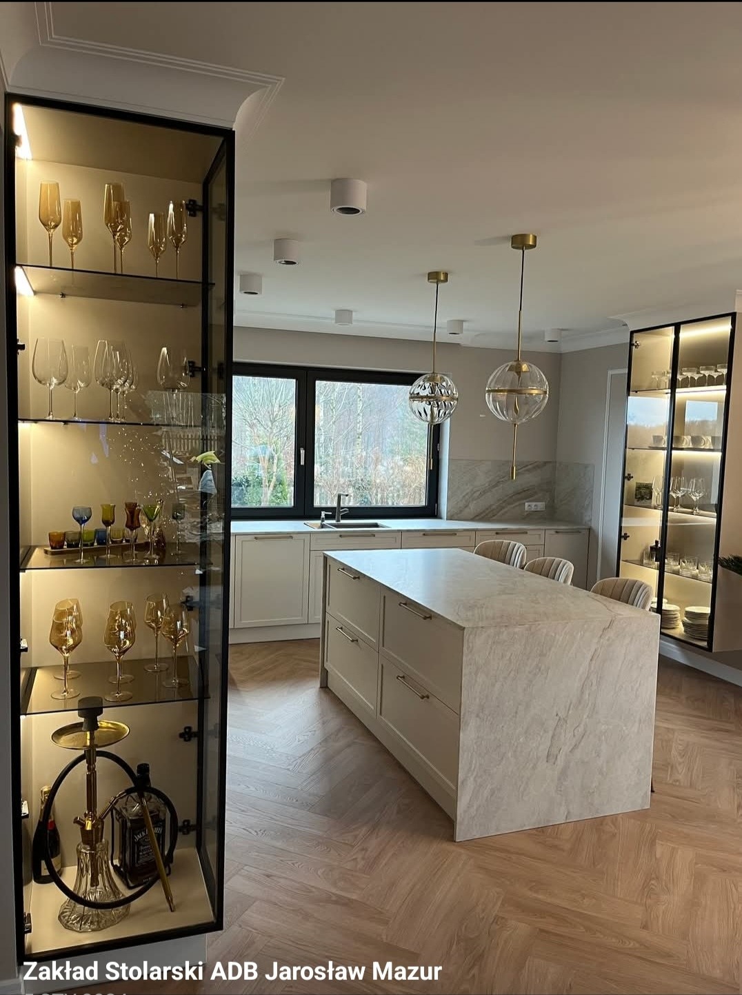 Jasna kuchnia z wyspą i przeszklonymi szafkami z podświetleniem LED. Na półkach szkło dekoracyjne i akcesoria. Parkiet ułożony w jodełkę francuską.
