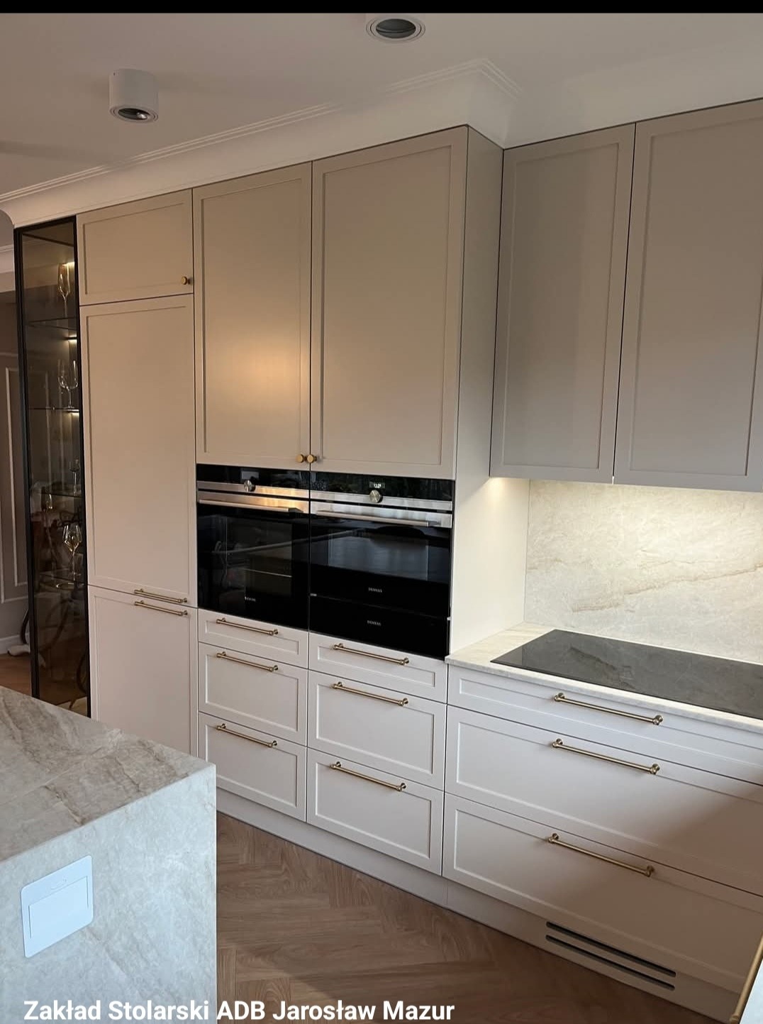 Jasna, elegancka kuchnia na wymiar z frezowanymi frontami, złotymi uchwytami i zabudowanym sprzętem AGD. Widoczna przeszklona witryna.