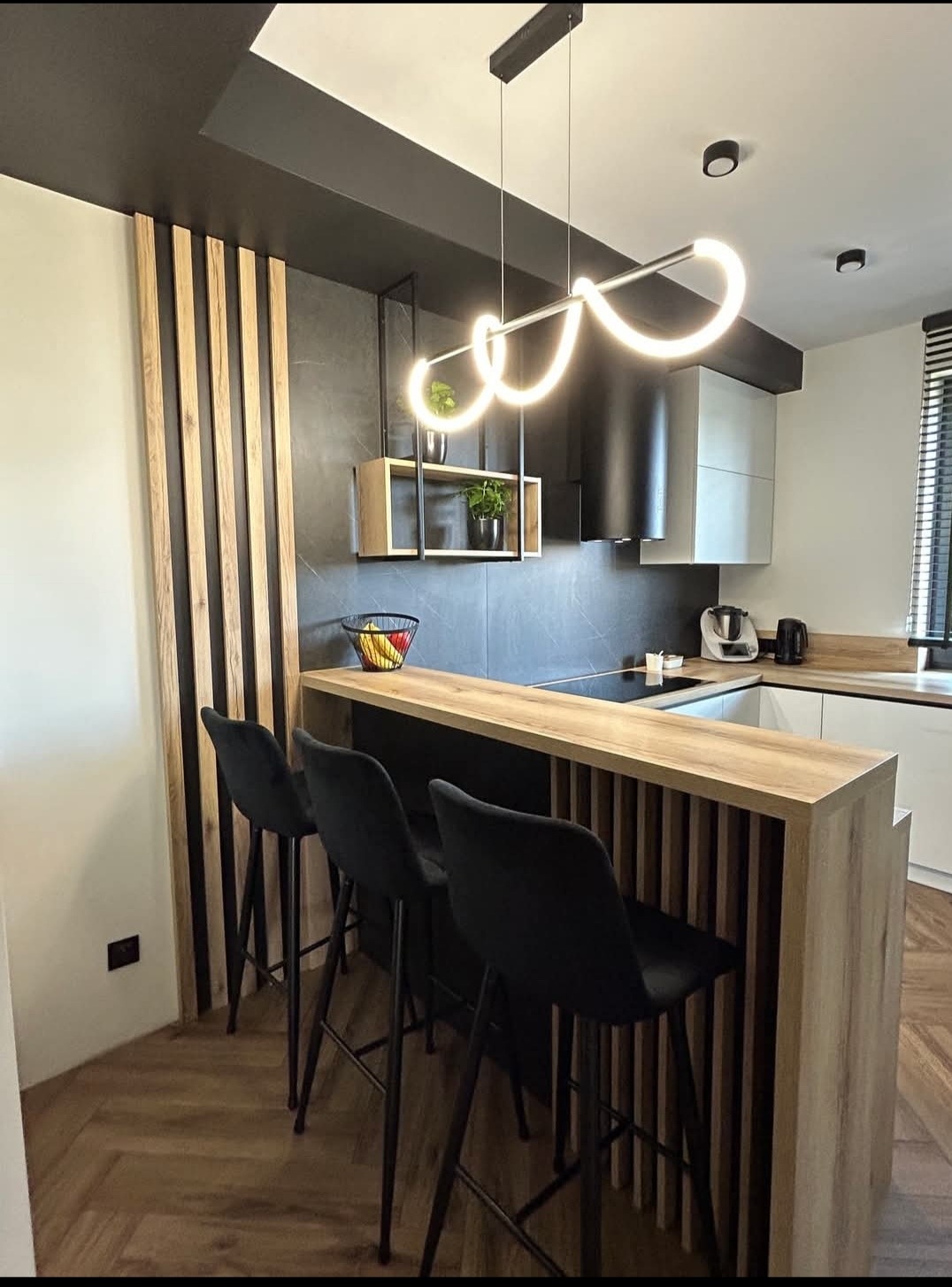 Nowoczesna kuchnia z wyspą i hokerami, oświetleniem LED i drewnianymi lamelami. Czarne i białe fronty, drewniany blat i podłoga w jodełkę. Minimalistyczny design.
