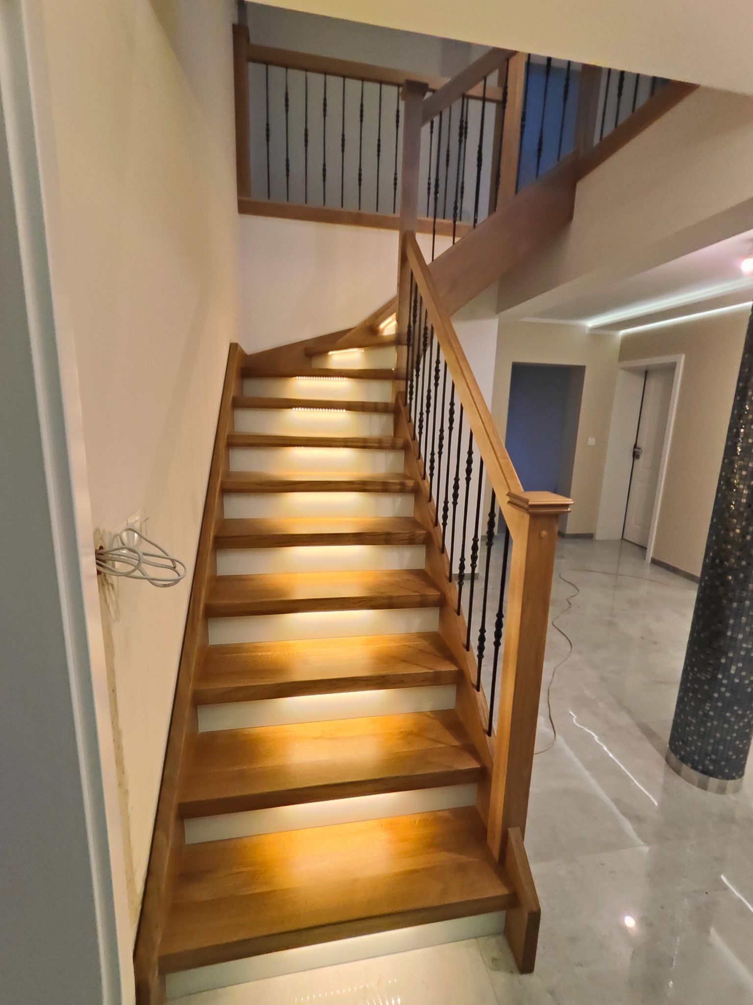 Klatka schodowa z drewnianymi stopniami podświetlonymi od spodu, czarne metalowe balustrady, widok z dołu, marmurowe podłoże i fragment wystającej instalacji.