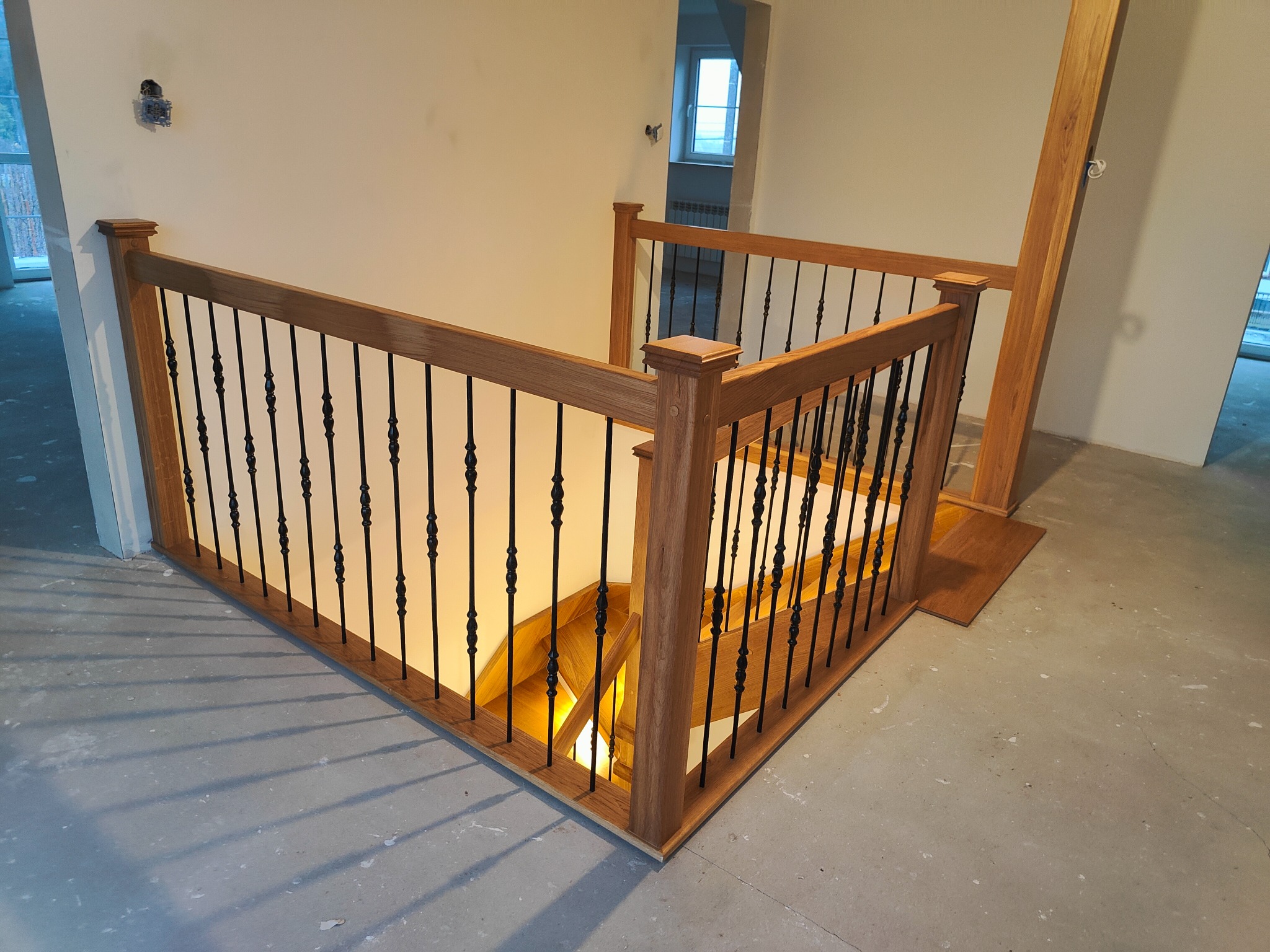 Drewniane schody z czarnymi, kutymi balustradami, widoczne z góry. Naturalne oświetlenie podkreśla detale wykończenia. Surowa podłoga w tle.