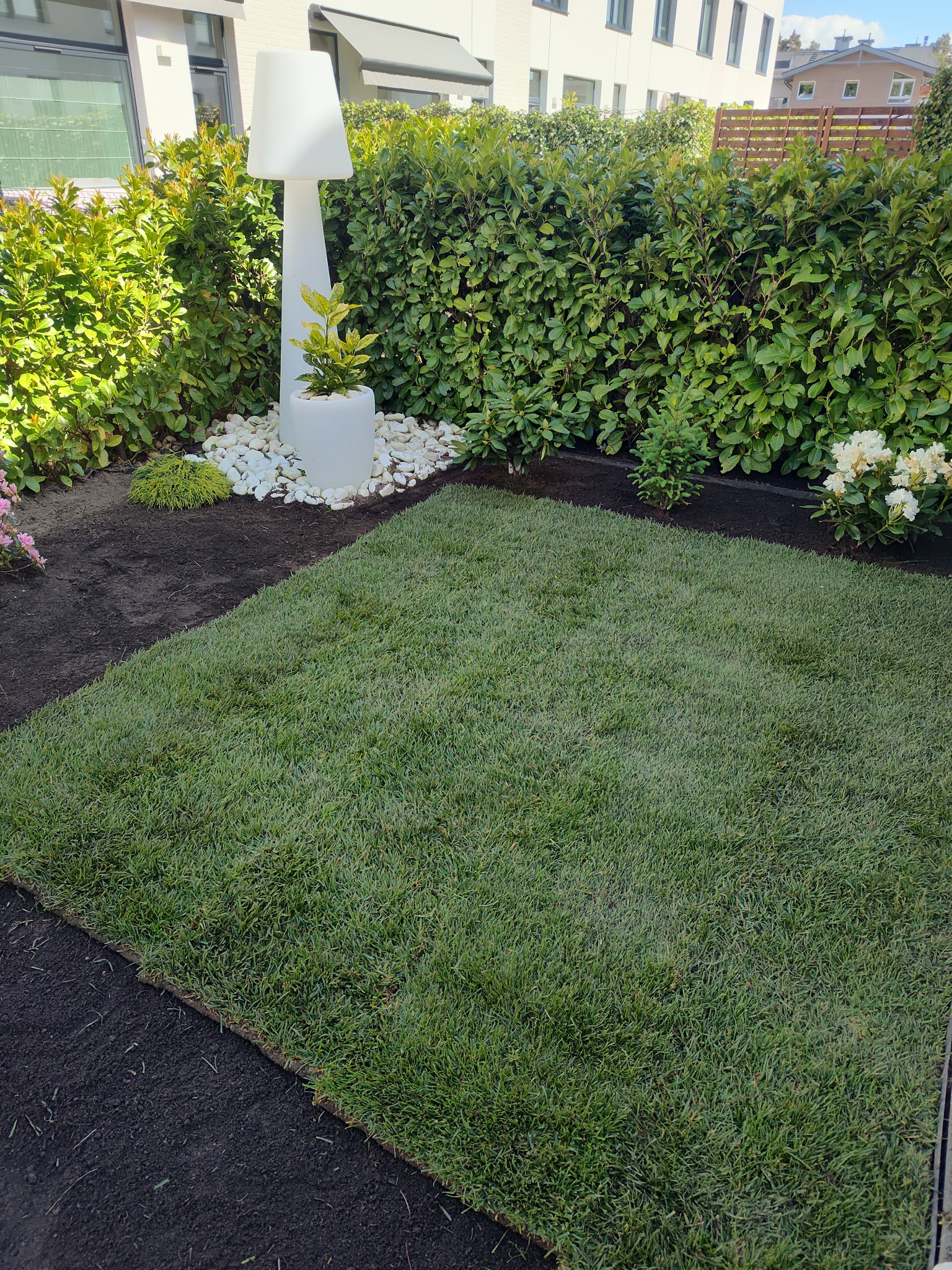 Fragment ogrodu z ułożoną trawą z rolki, obok rabata z ziemią, biała lampa ogrodowa i żywopłot. Widoczne elementy małej architektury ogrodowej.