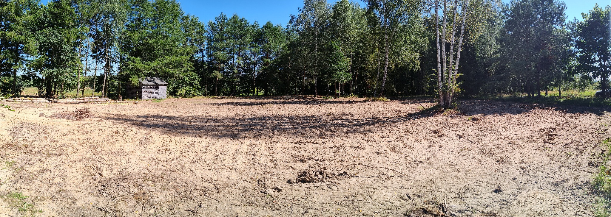 Panoramiczny widok na teren po niwelacji, z widocznymi pozostałościami po wycince drzew i małym drewnianym budynkiem w tle, przy słonecznej pogodzie.