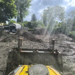 Widok z kabiny żółtej koparki na teren budowy z hałdami ziemi, drzewami i budynkiem w tle, w słoneczny dzień.