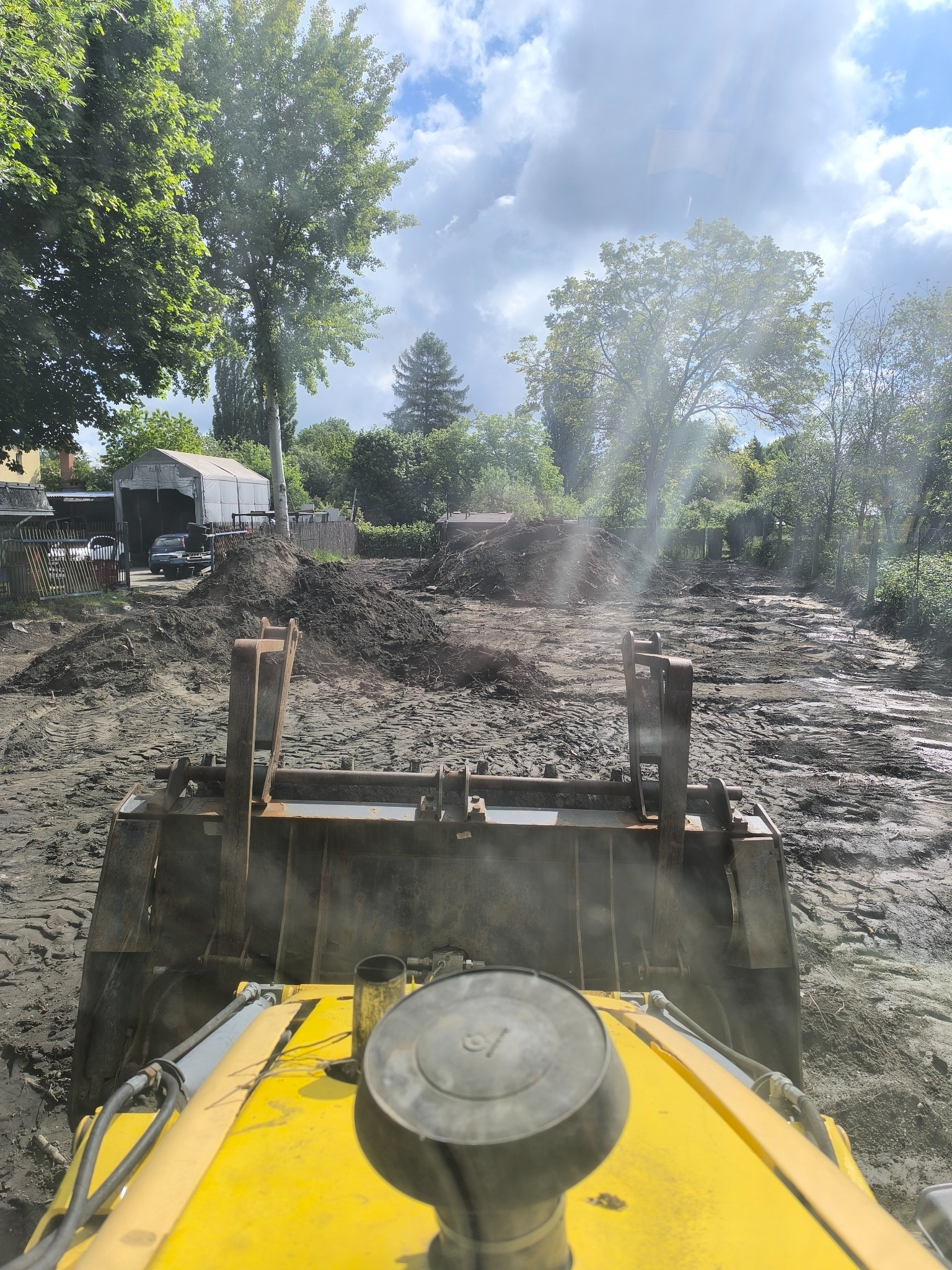 Widok z kabiny żółtej koparki na teren budowy z hałdami ziemi, drzewami i budynkiem w tle, w słoneczny dzień.