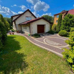 PGCLEAN - Zadbany ogród z kostką brukową i trawnikiem przed domem z garażem. Widoczne drzewa iglaste i liściaste, niebieskie niebo z chmurami. Wąż ogrodowy leży na trawie.