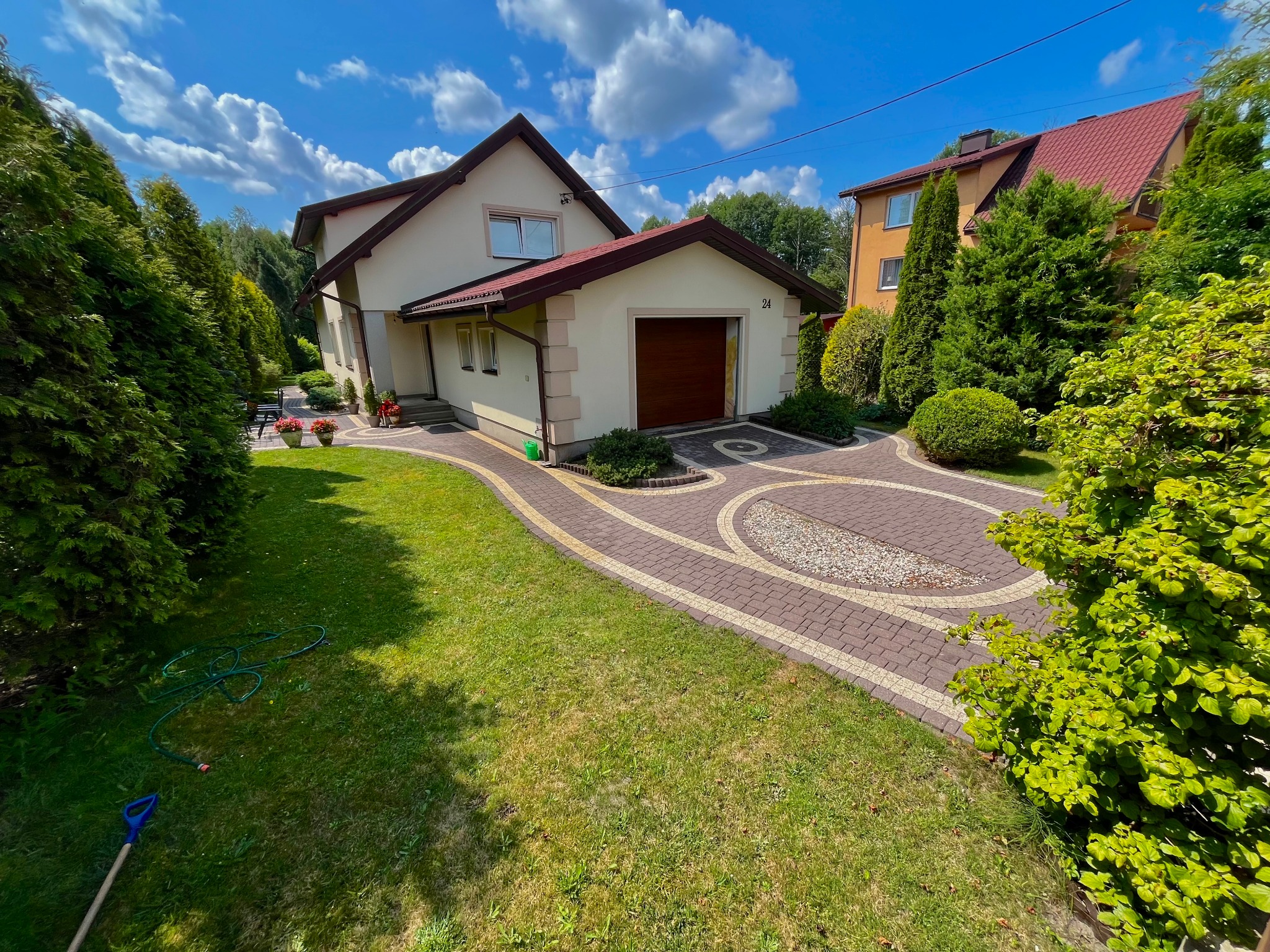 Zadbany ogród z kostką brukową i trawnikiem przed domem z garażem. Widoczne drzewa iglaste i liściaste, niebieskie niebo z chmurami. Wąż ogrodowy leży na trawie.
