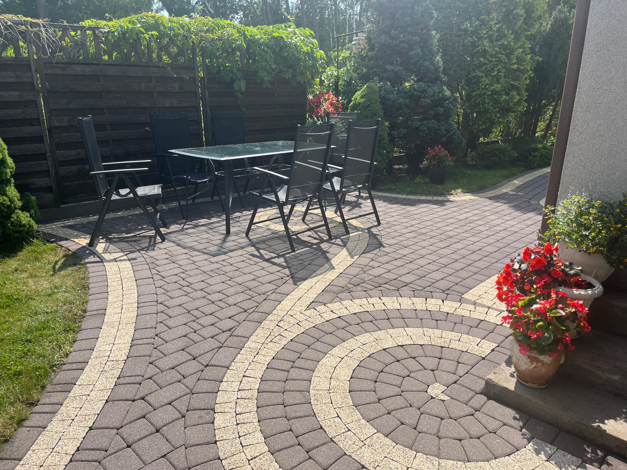 Ułożona kostka brukowa z dekoracyjnym wzorem na posesji w Łochowie. Widoczne meble ogrodowe, rośliny i elementy architektury.