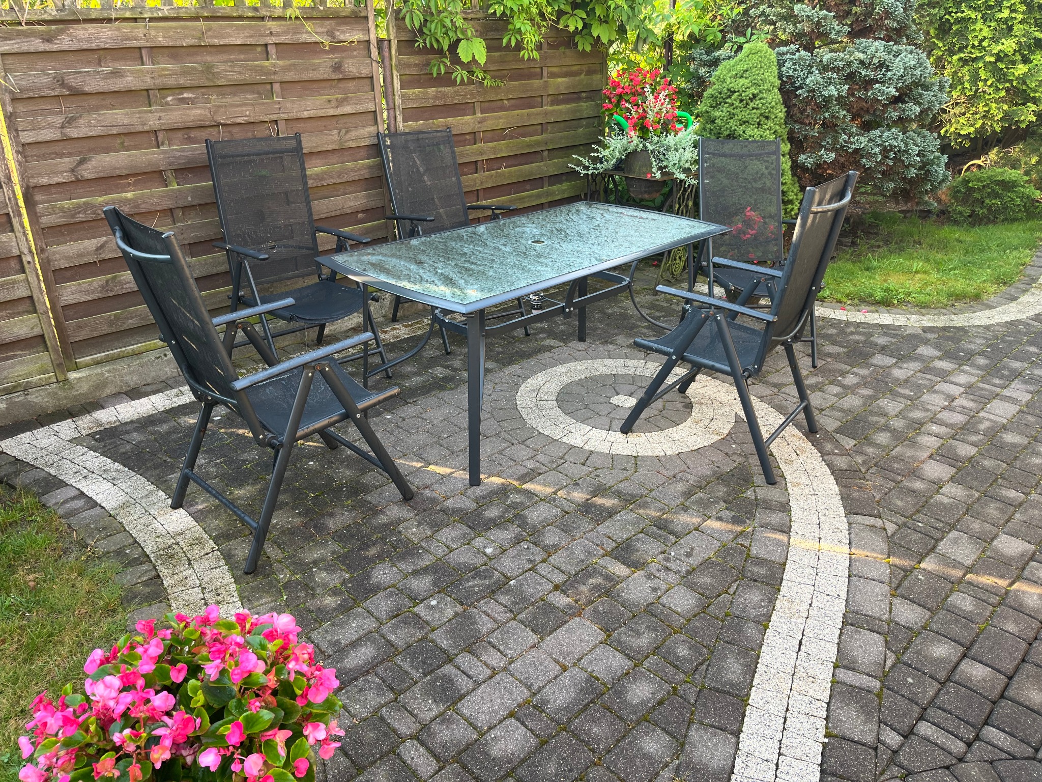 Zestaw mebli ogrodowych (stół i krzesła) na kostce brukowej z delikatnym porostem mchu, otoczony zielenią i kwiatami, przy drewnianym płocie.