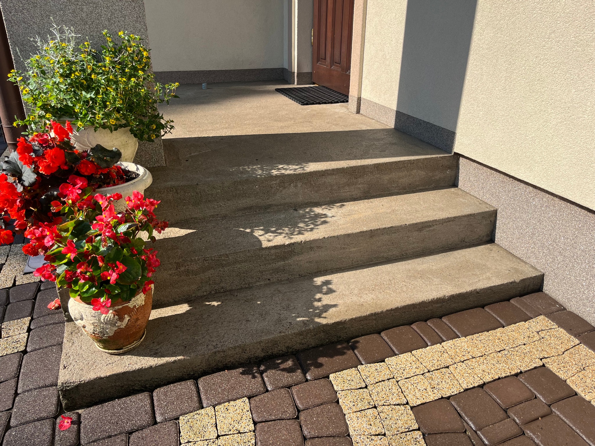 Schody wejściowe z kostki brukowej i kwiatami w donicach, prowadzące do drzwi wejściowych. Cień drzewa na schodach. Słoneczny dzień.