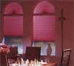Dwa okna z plisowanymi roletami w odcieniach różu, częściowo zasłonięte, widoczne elementy wystroju wnętrza, w tym dekoracyjny wazon i kwiaty na stole.