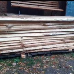 Stos tarcicy z jasnego drewna, zabezpieczony metalowymi taśmami, leżący na drewnianym podłożu na zewnątrz. W tle ławka i ceglany budynek.