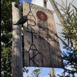 Oskar Steleblak - Tablica informacyjna z napisem 'Tartak' i numerem telefonu, umieszczona na drewnianym słupie w otoczeniu zieleni. Widoczna tarcza piły jako element dekoracyjny.